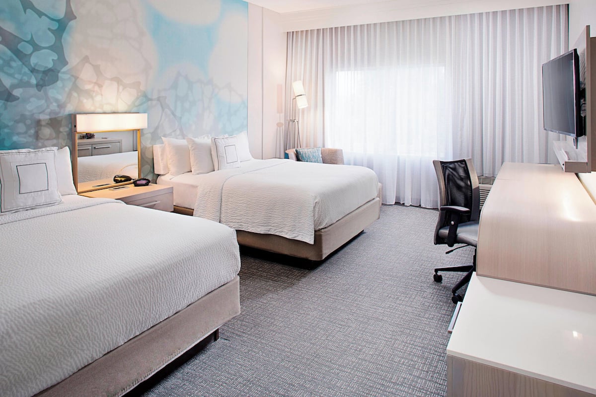 Ein Hotelzimmer mit zwei Betten, einem Schreibtisch, einem Stuhl, einem Fernseher und einem großen Fenster mit Vorhängen.