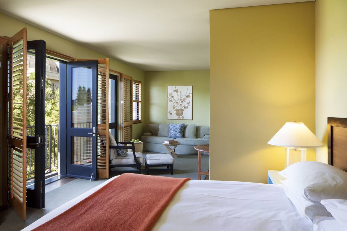 A hotel room with a bed, a sofa, a chair, a table, and a lamp. There is a door leading to a balcony.