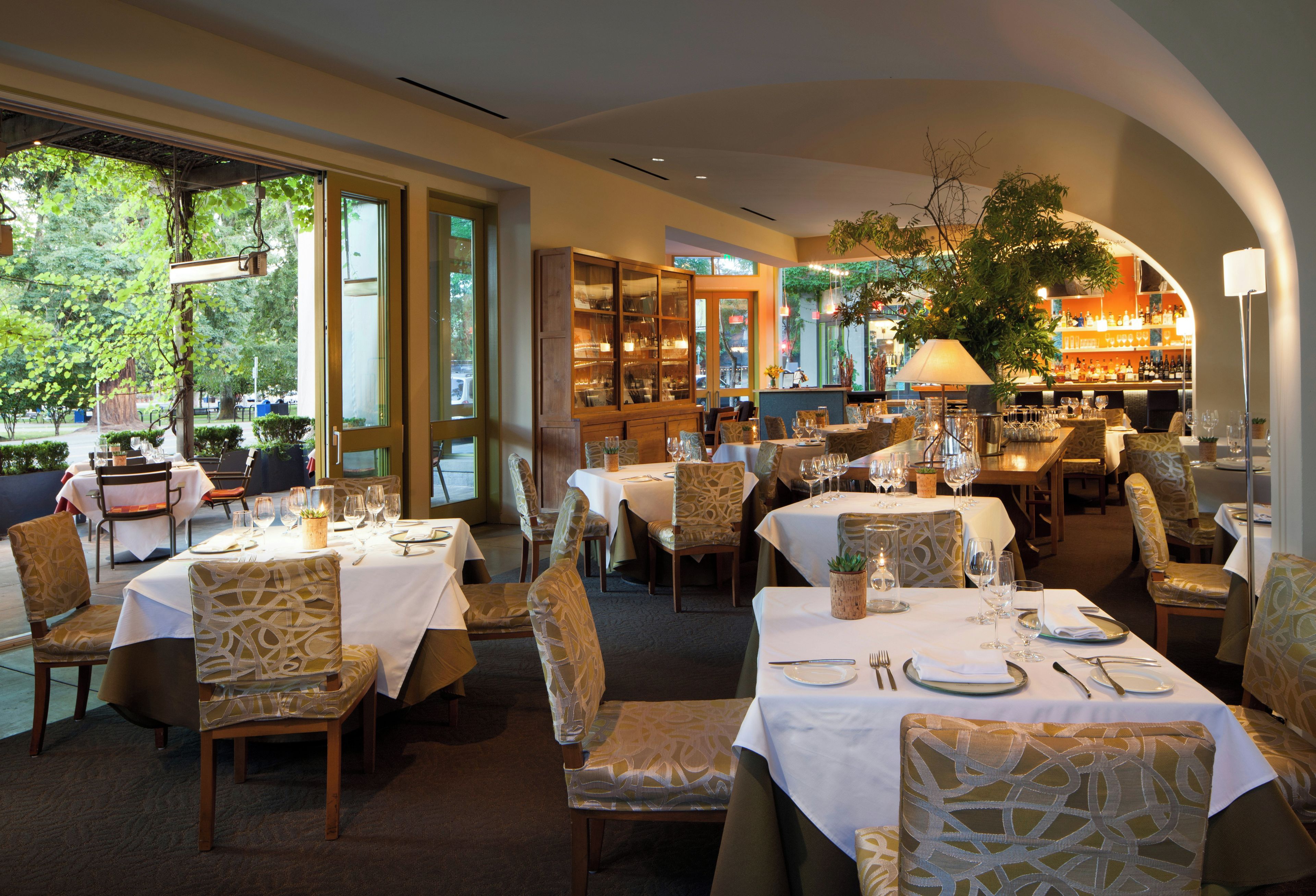 Restaurante com mesas preparadas para refeições, cadeiras e área de bar com garrafas e copos.
