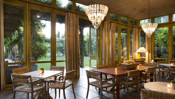 Un comedor con sillas y mesas de mimbre, ventanas amplias con cortinas y un candelabro.