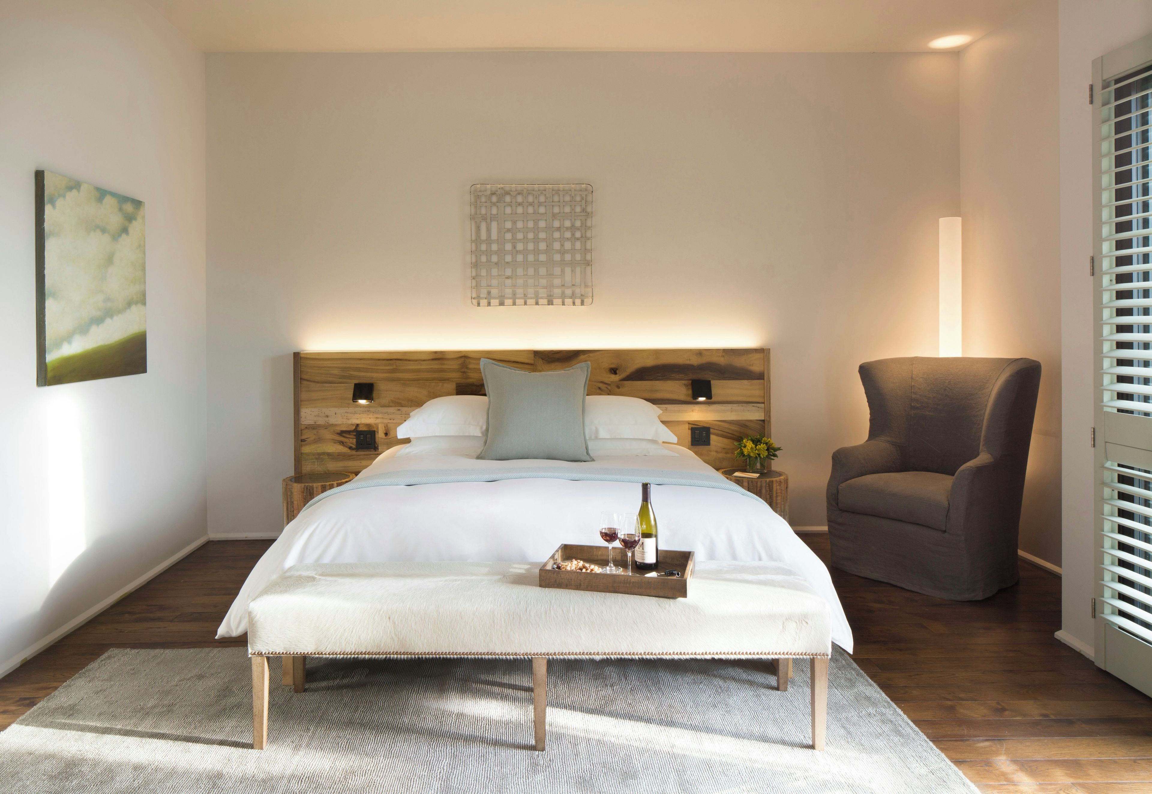 A bedroom with a bed, bedside tables, a chair, a tray with wine glasses and a bottle, and a painting on the wall.