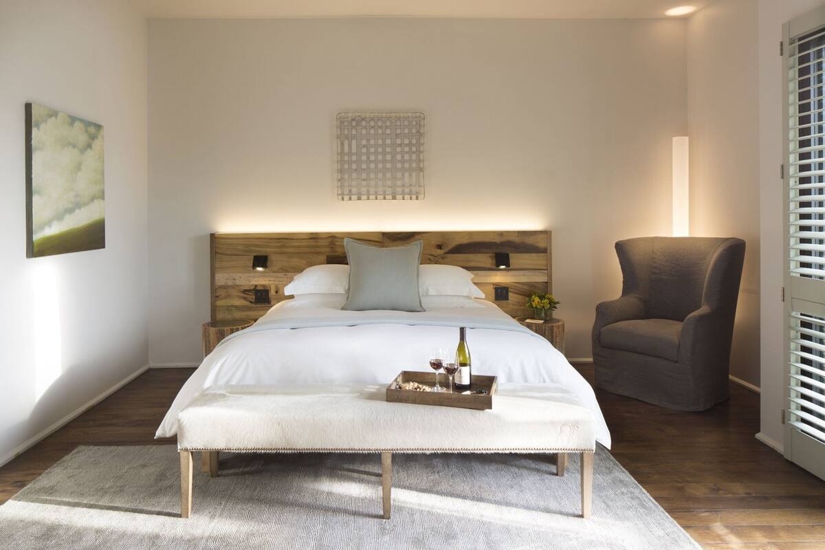 Un dormitorio con una cama grande, una mesita de noche con lámpara, una silla y una ventana con persianas.