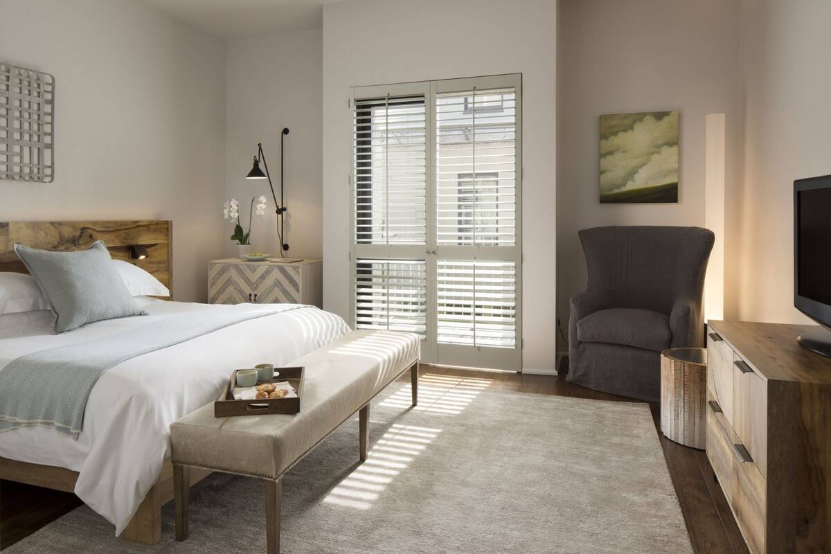 A bedroom with a bed, bench, bedside table, lamp, chair, TV stand, and window blinds.
