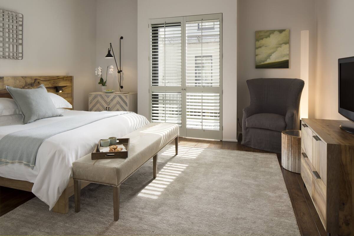 A bedroom with a bed, bench, bedside table, lamp, chair, TV stand, and a window with blinds.