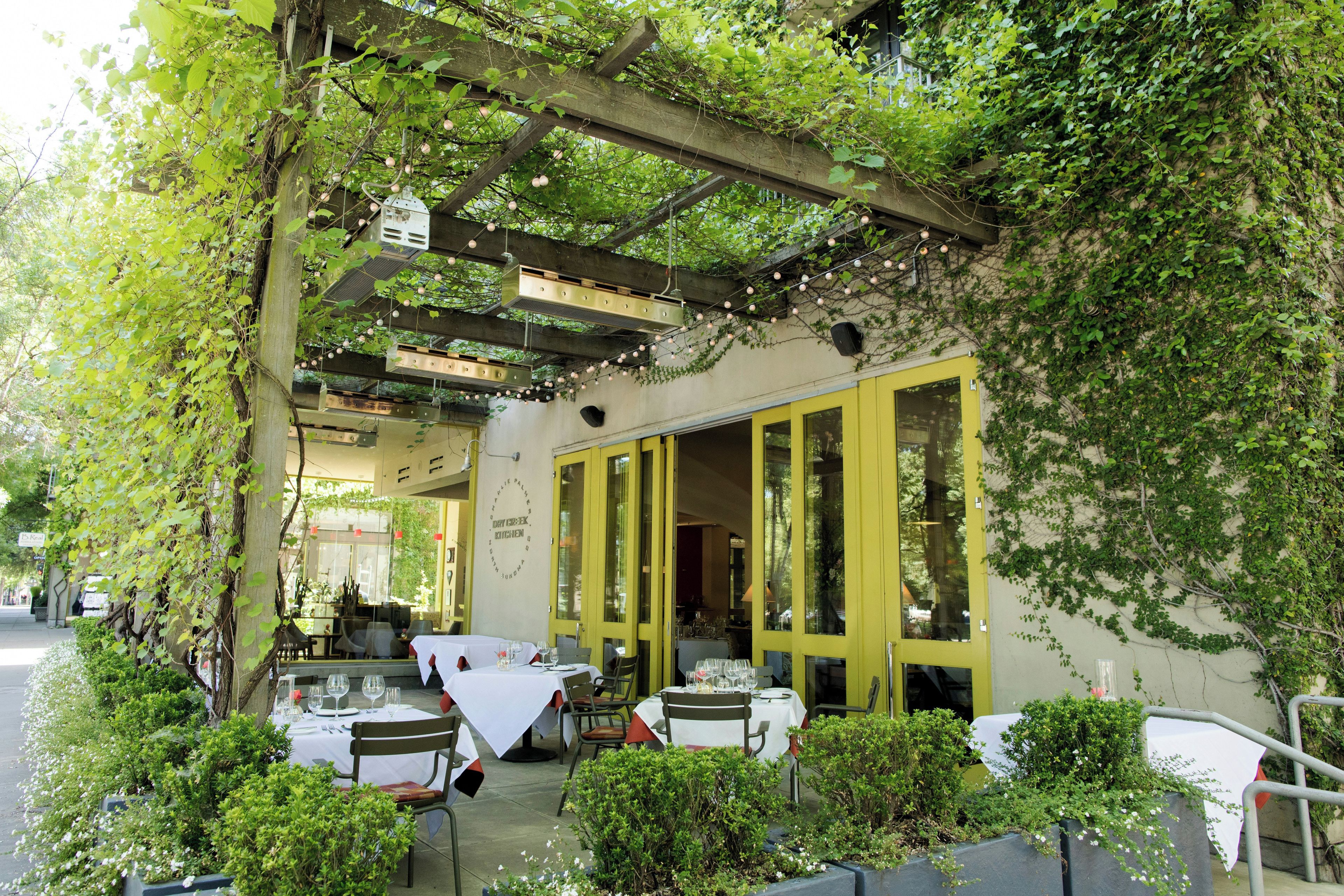 An outdoor dining area with tables and chairs, covered by a wooden pergola with hanging lights, and surrounded by greenery.