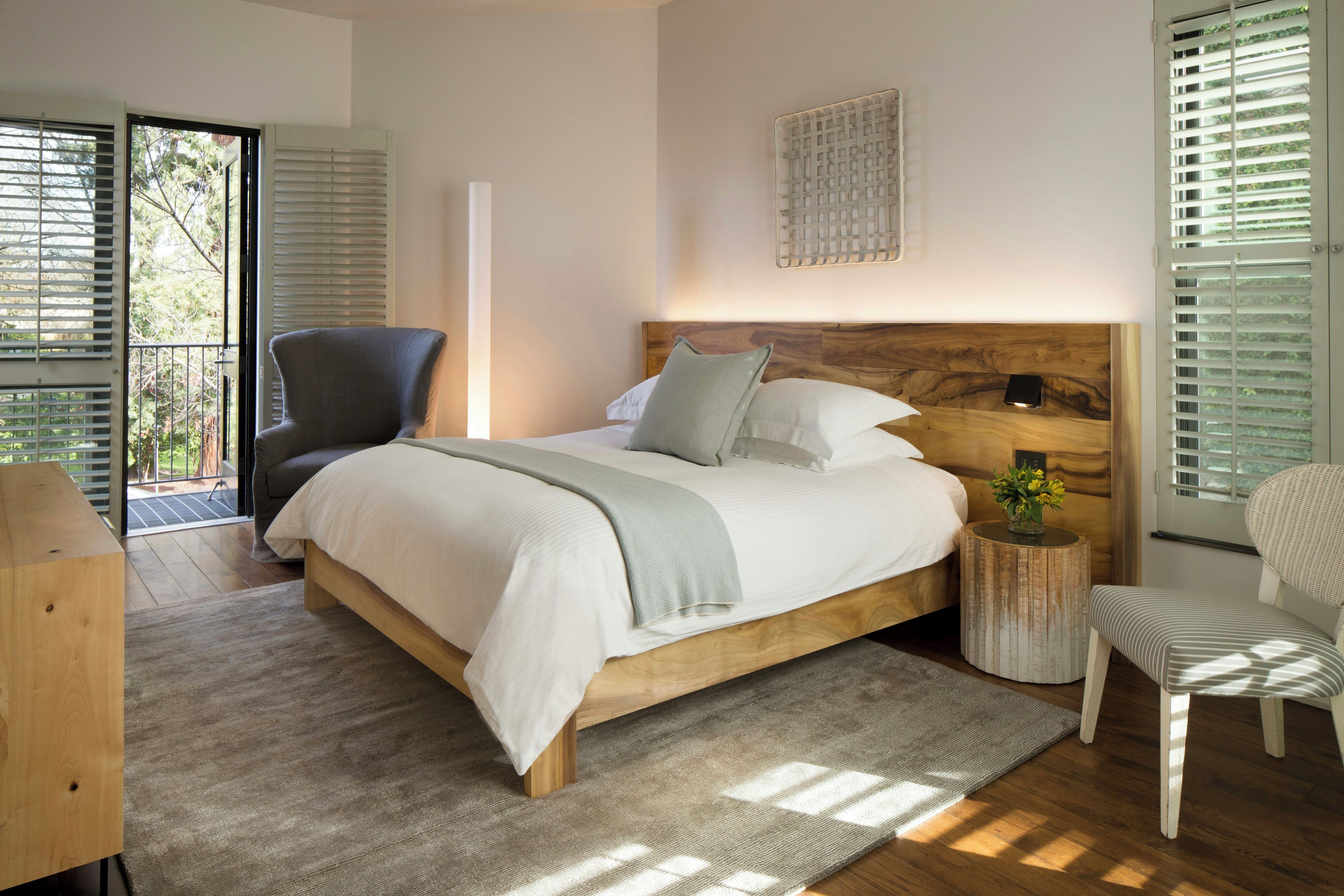 A bedroom with a wooden bed, a grey armchair, a round wooden side table with a vase, and a view of the outdoors through the window.