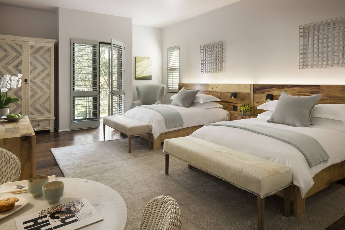 A modern bedroom with two beds, a wooden headboard, a round table with a magazine, and a view of the outdoors through a window.
