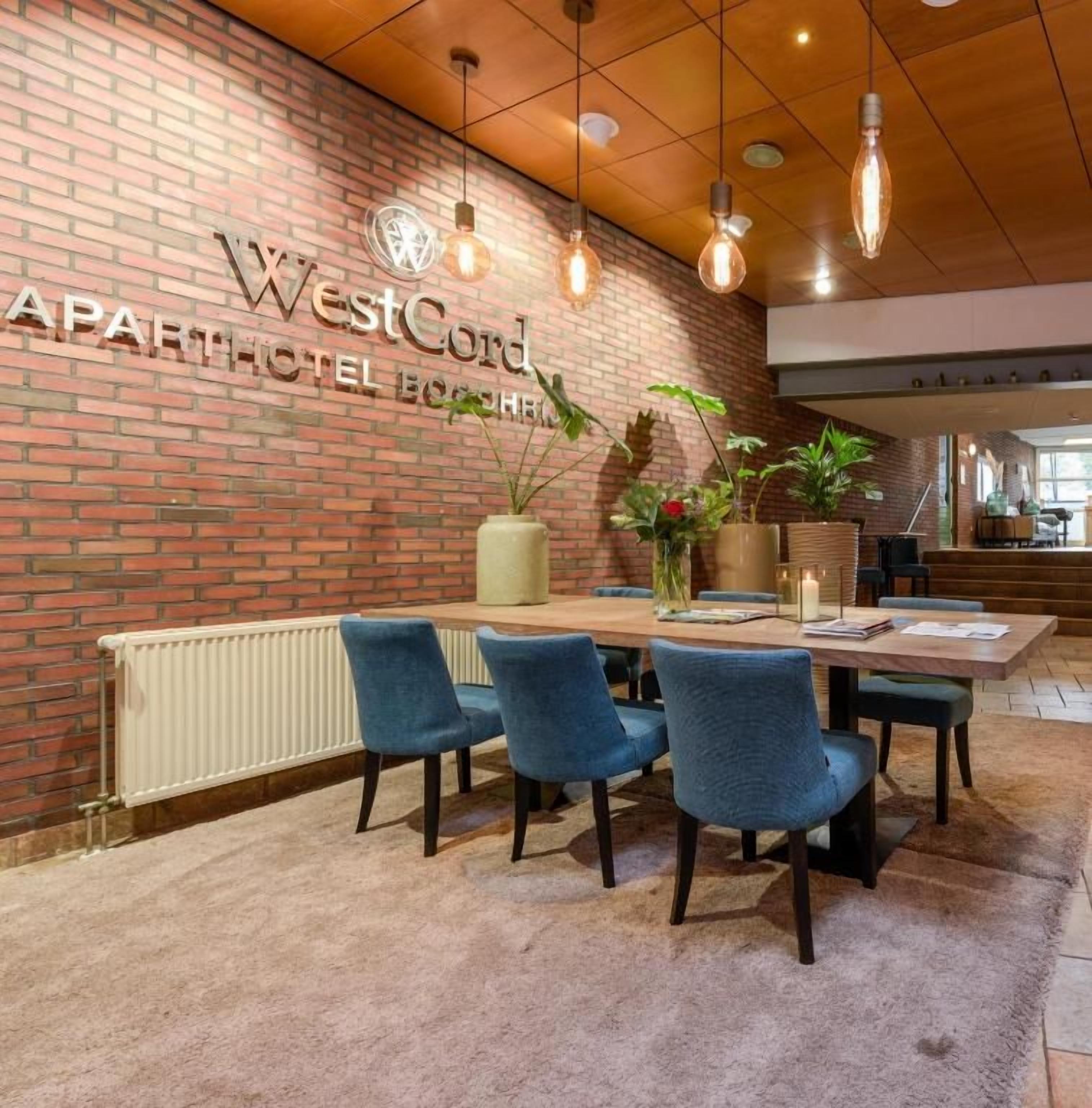 Ein Innenraum eines WestCord Apartmenthotels mit Backsteinwand, einem Essbereich mit blauen Stühlen und hängenden Leuchten.