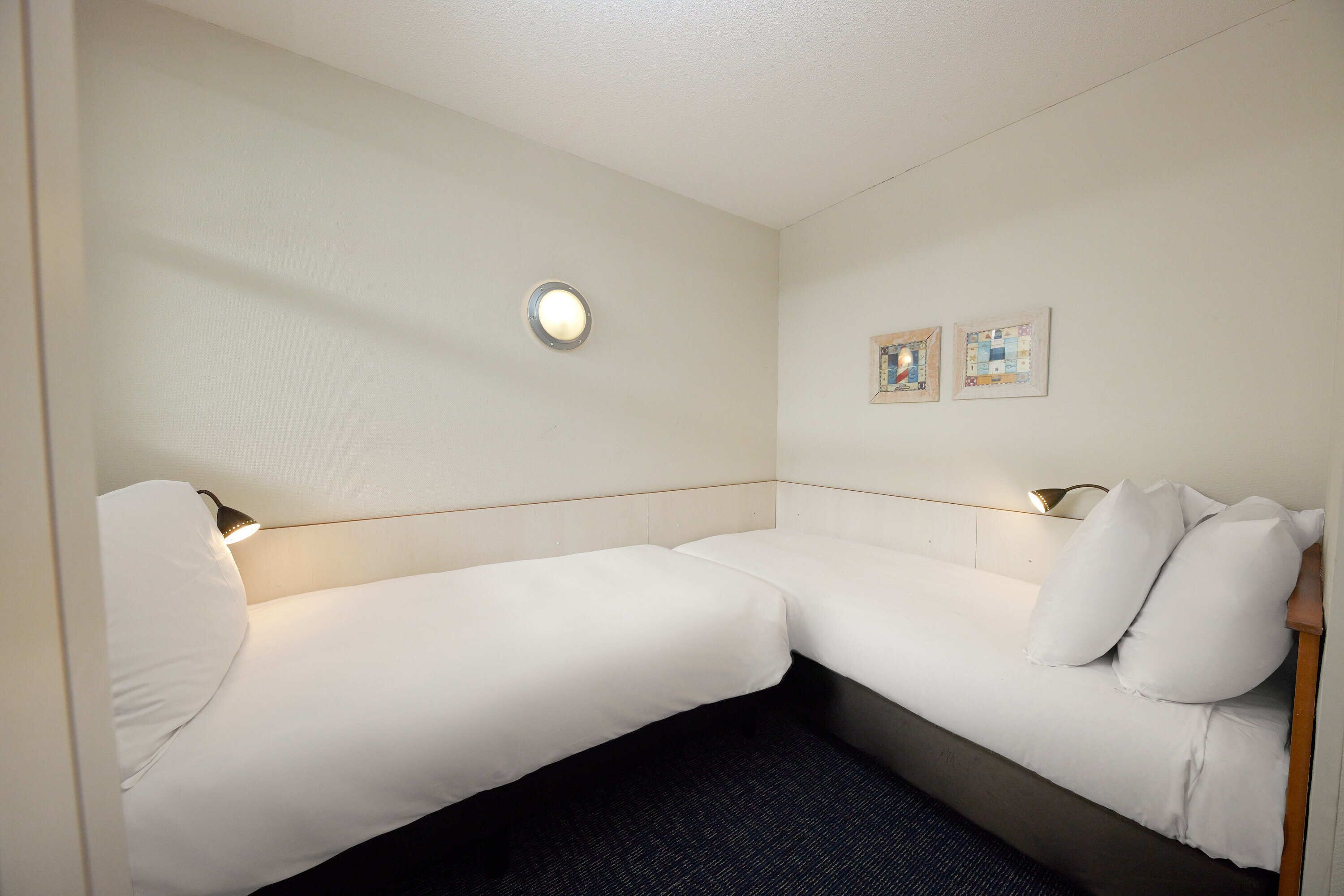 Habitación de hotel con dos camas individuales, una lámpara de pared redonda y dos cuadros enmarcados.