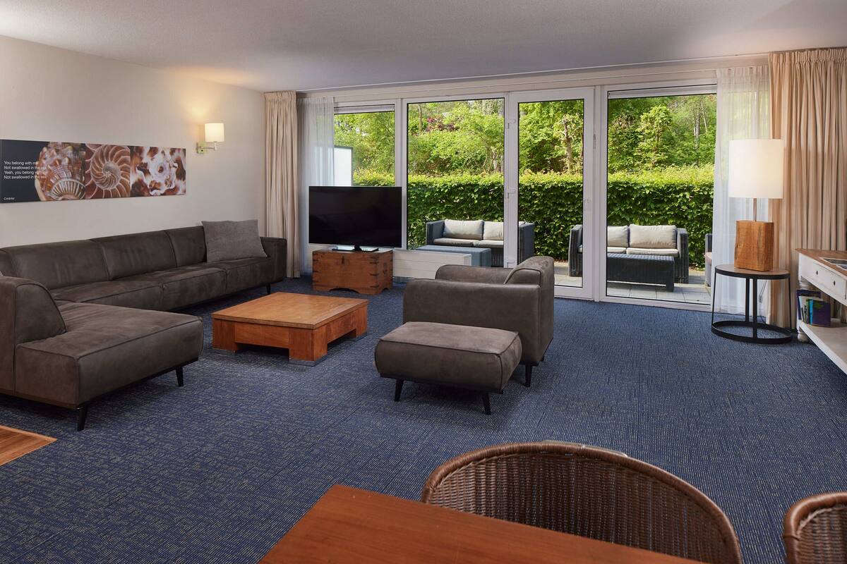 Una habitación de hotel moderna con un sofá grande, un televisor, una mesa de centro de madera y una silla de mimbre.