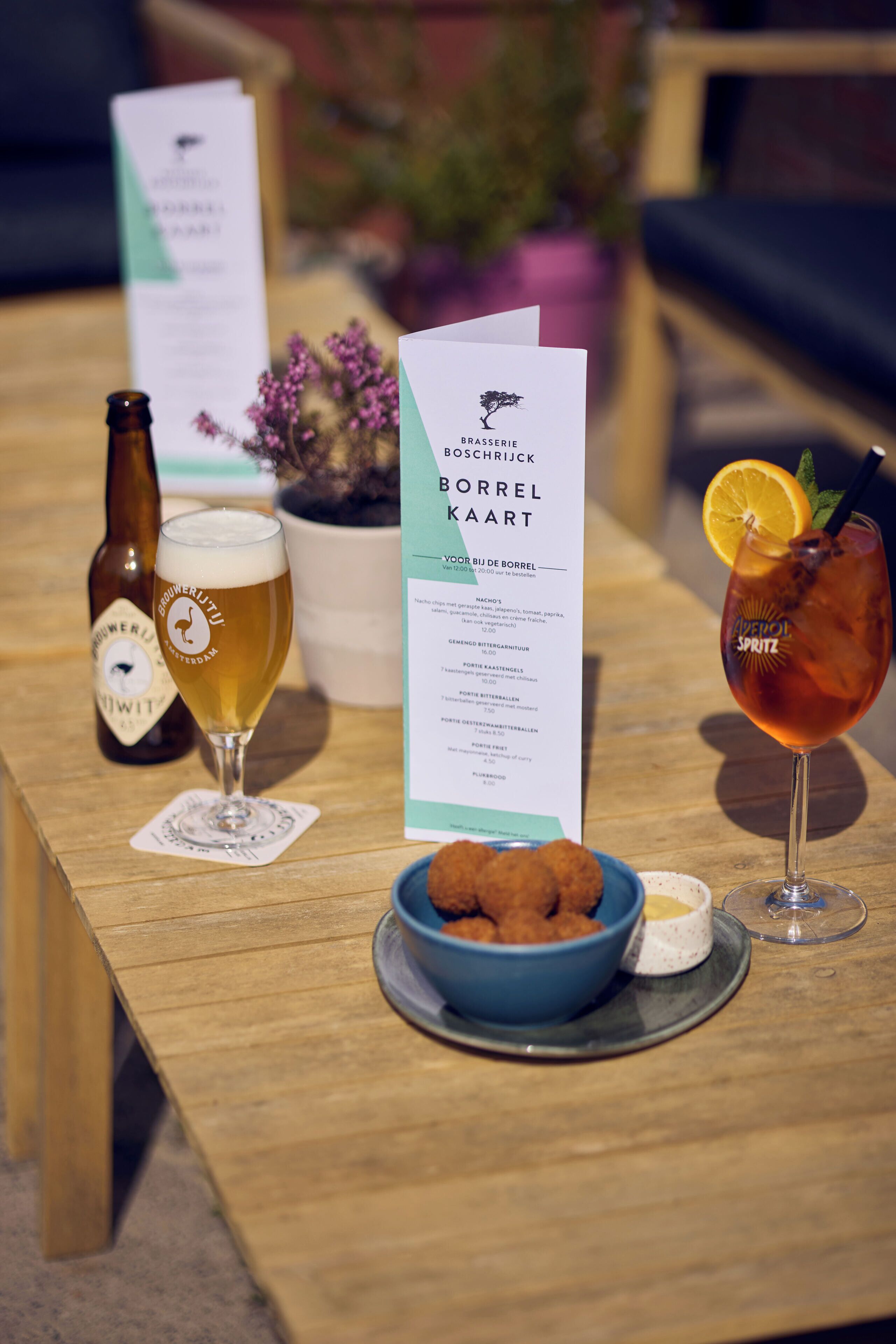 A wooden table with a bottle of beer, a glass of beer, a menu, a bowl of snacks, and a cocktail glass with an orange slice.