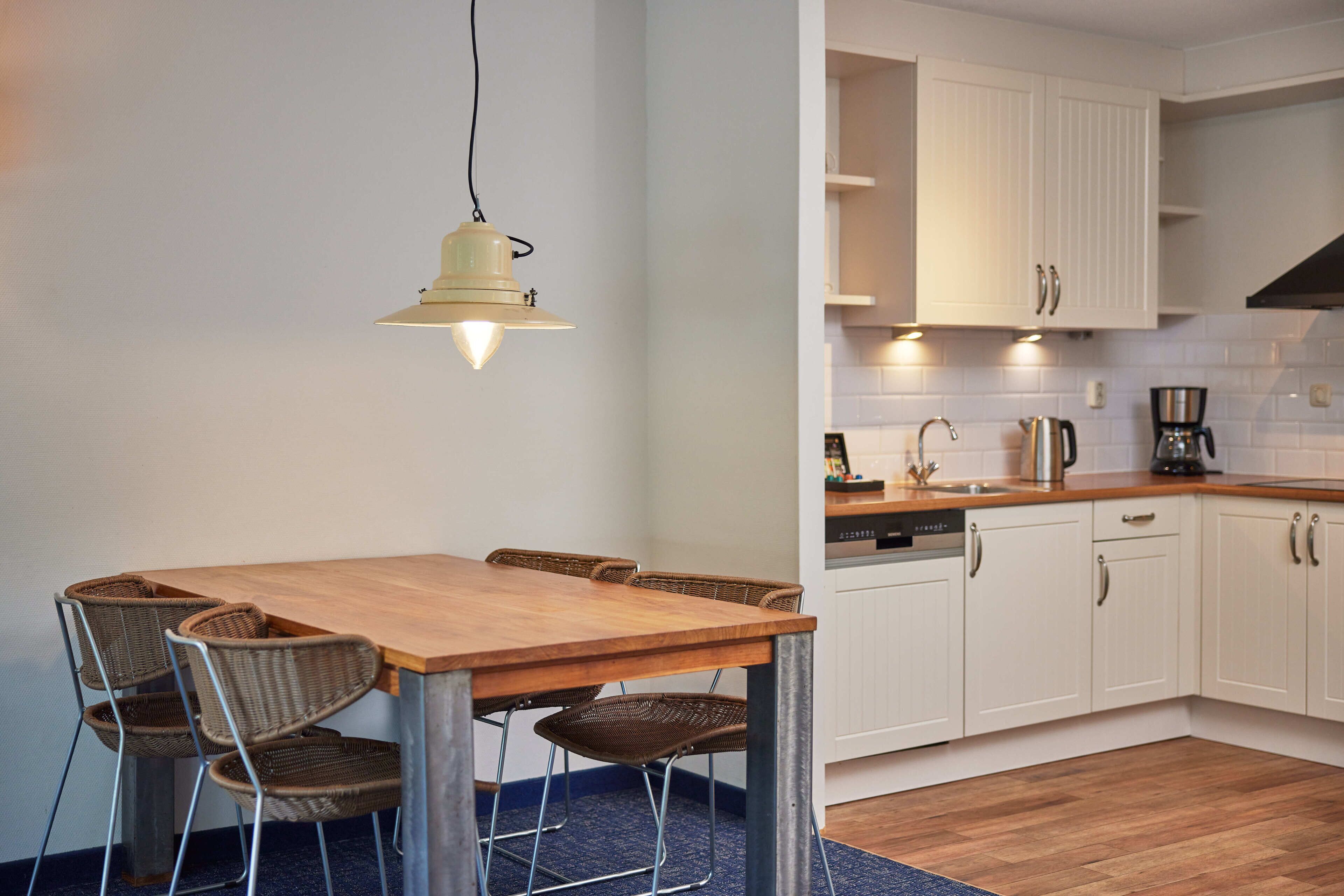 A dining area with a wooden table and wicker chairs, a hanging light fixture, and white cabinetry in the adjacent kitchen area.
