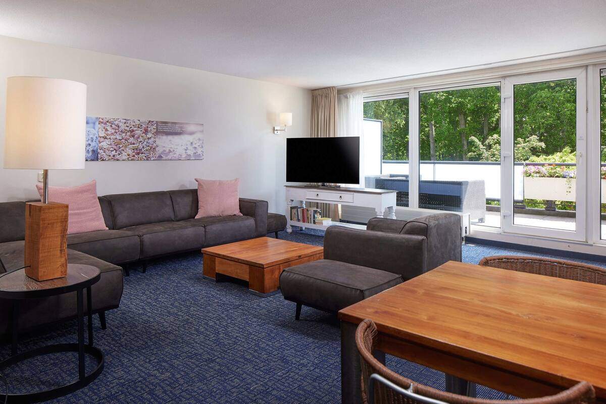 Habitación de hotel moderna con sofá gris, una mesa de centro de madera, televisión y un ventanal con vistas a zonas verdes.