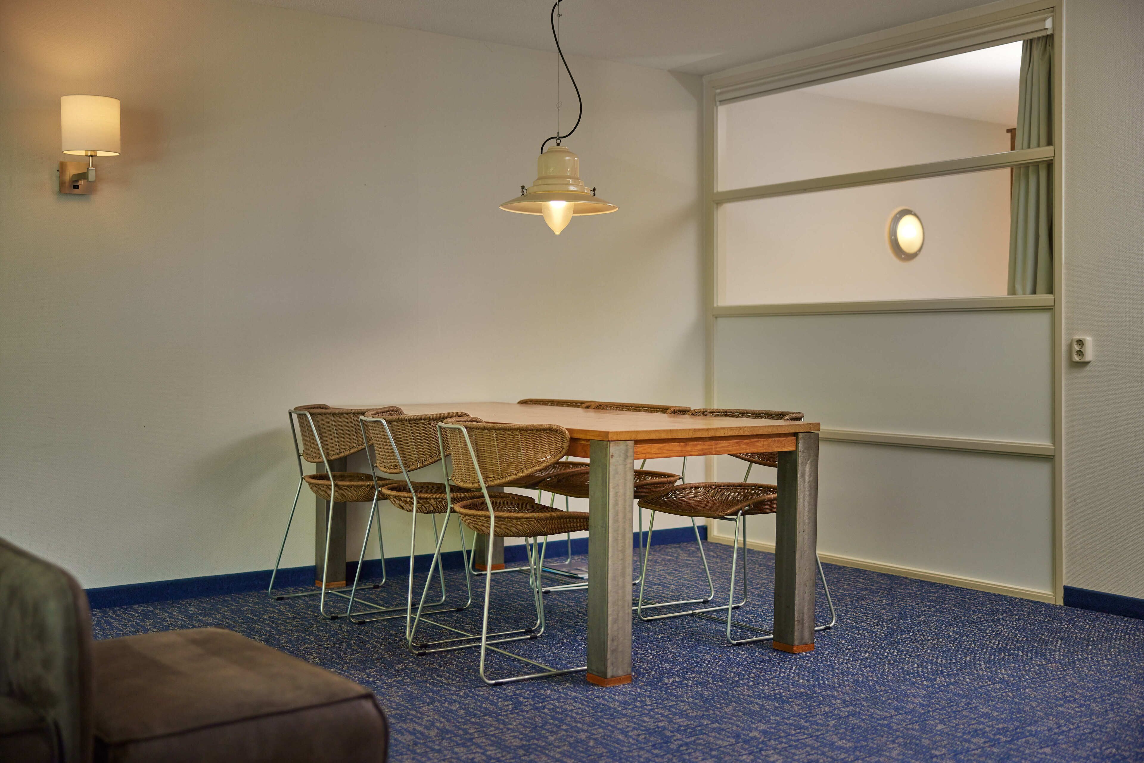 Une pièce comprenant une table à manger, quatre chaises en osier, un luminaire suspendu et une porte dotée d’un panneau en verre dépoli.