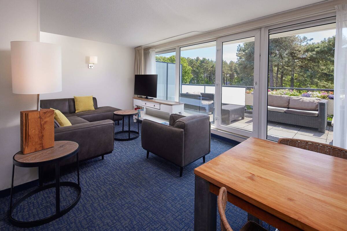 Habitación de hotel con sofá, mesa de madera, televisión y balcón con mobiliario exterior.