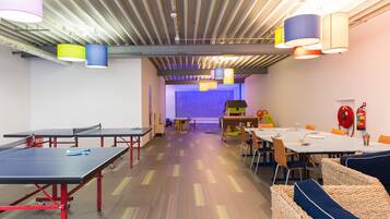Una habitació espaiosa amb taules de ping-pong, un escenari amb un fons de color i un extintor de foc a la paret.