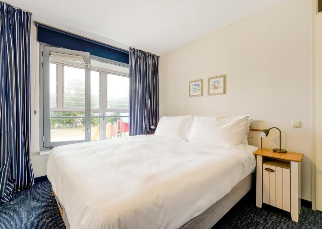 Habitación de hotel con una cama grande, una mesita de noche con lámpara y una ventana con cortinas.
