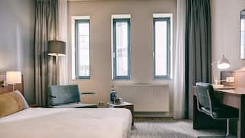 Ein Hotelzimmer mit einem Bett, zwei Sesseln, einem Schreibtisch, einer Lampe und drei Fenstern.