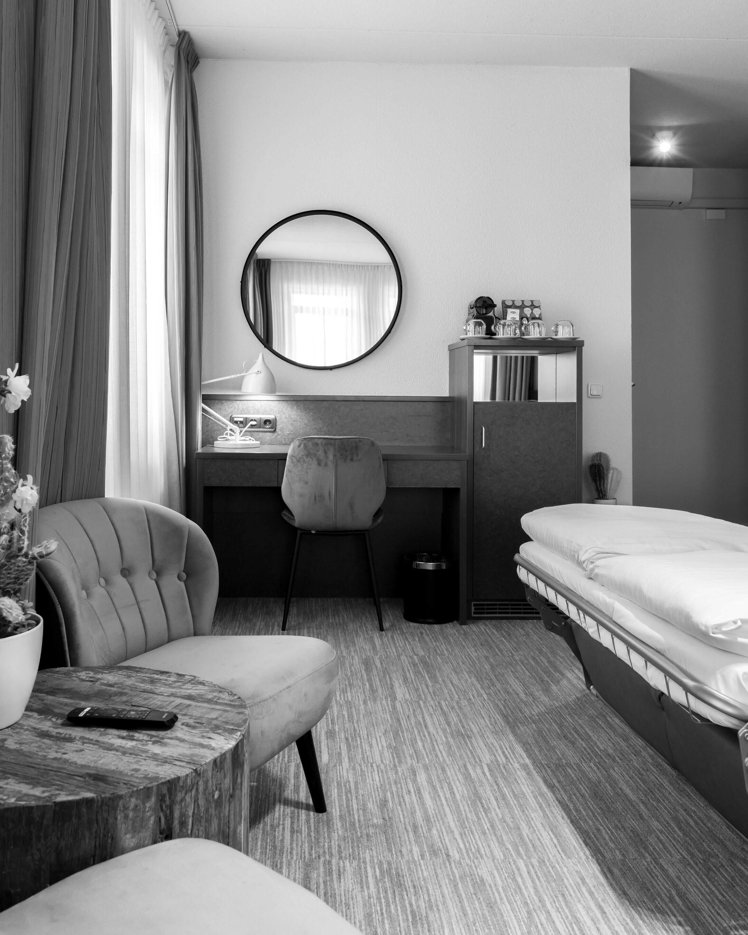 Een moderne hotelkamer met een bed, een bureau met een stoel, een ronde spiegel en een klein tafeltje met een afstandsbediening.