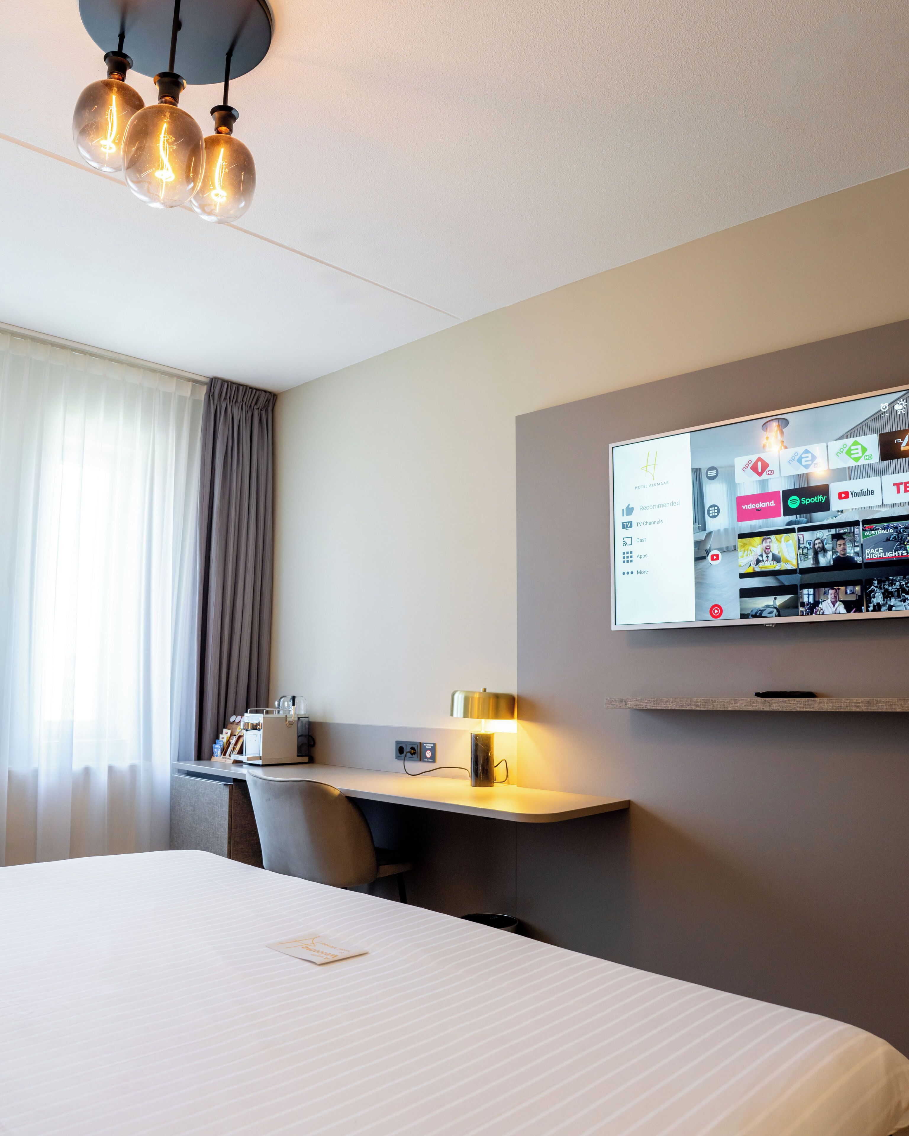 Ein Hotelzimmer mit Bett, Schreibtisch mit Lampe, Wandfernseher und einem Vorhangfenster.