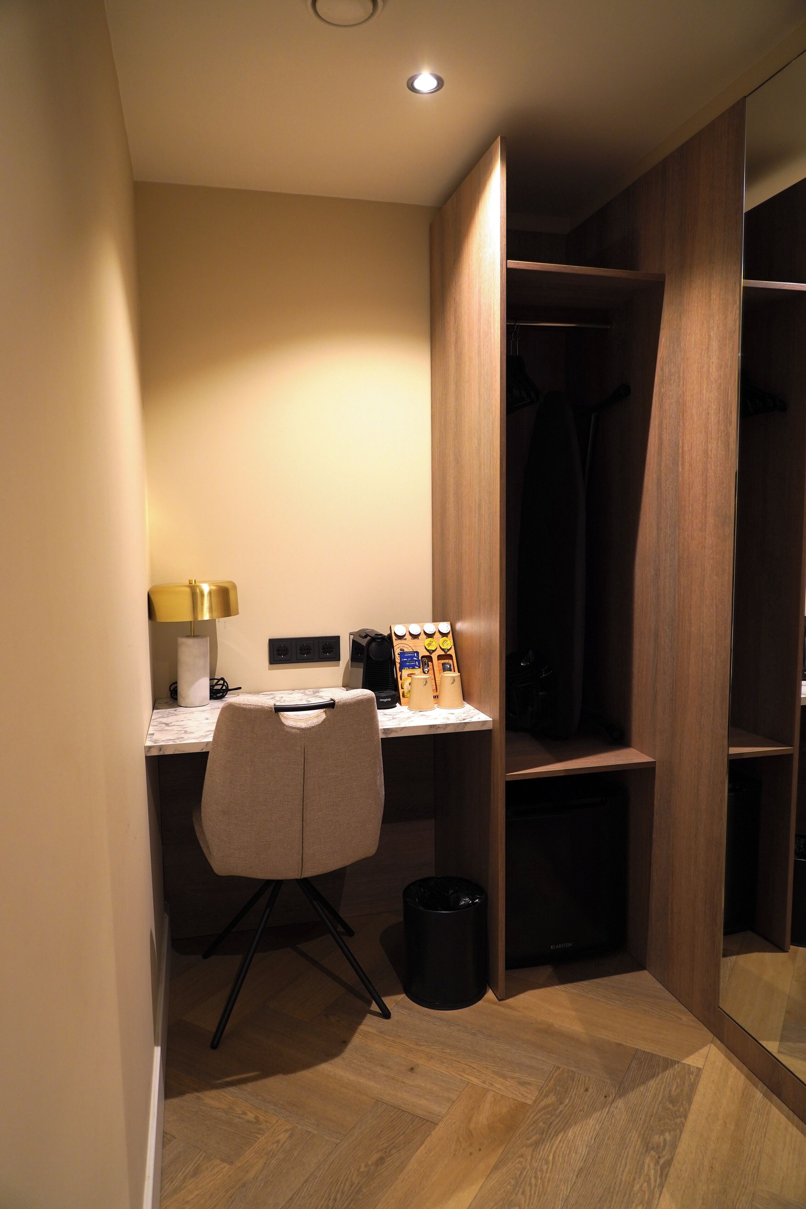 Une chambre d’hôtel de taille réduite, dotée d’un bureau en bois, d’une chaise, d’une petite poubelle et d’une armoire avec un miroir.