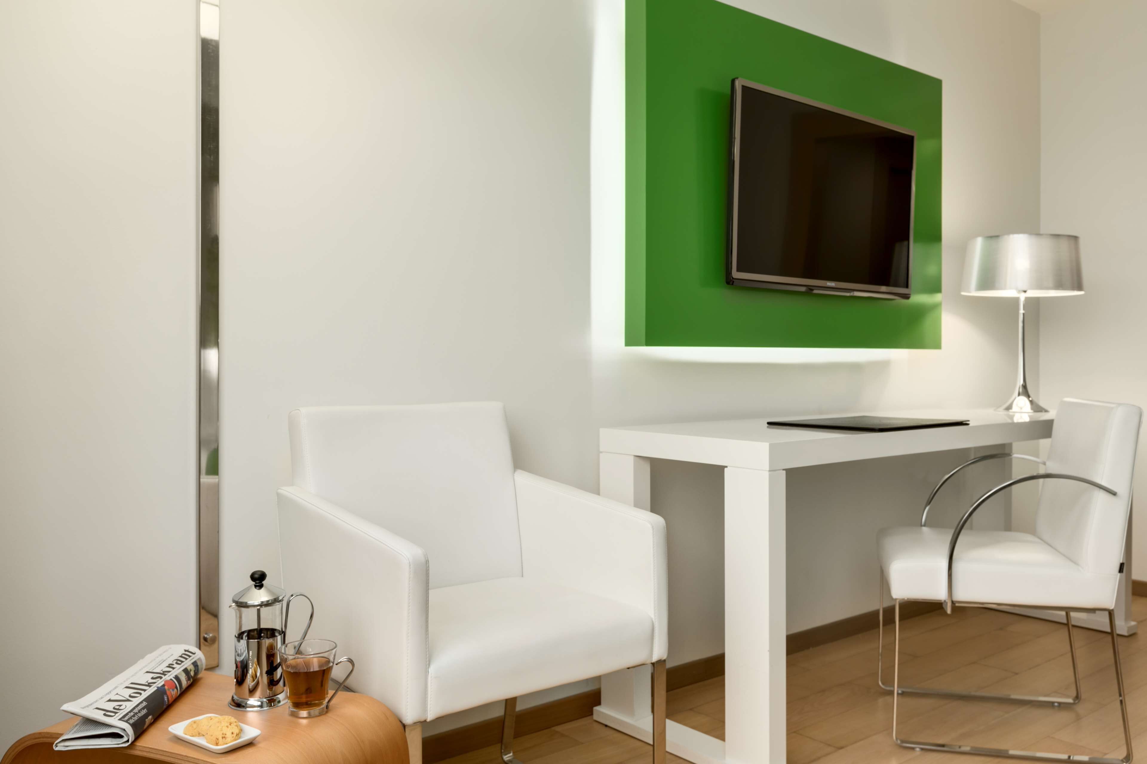 A modern hotel room with a white desk, two white chairs, a green accent wall, a flat-screen TV, a lamp, a newspaper, a coffee pot, and a cup of tea.