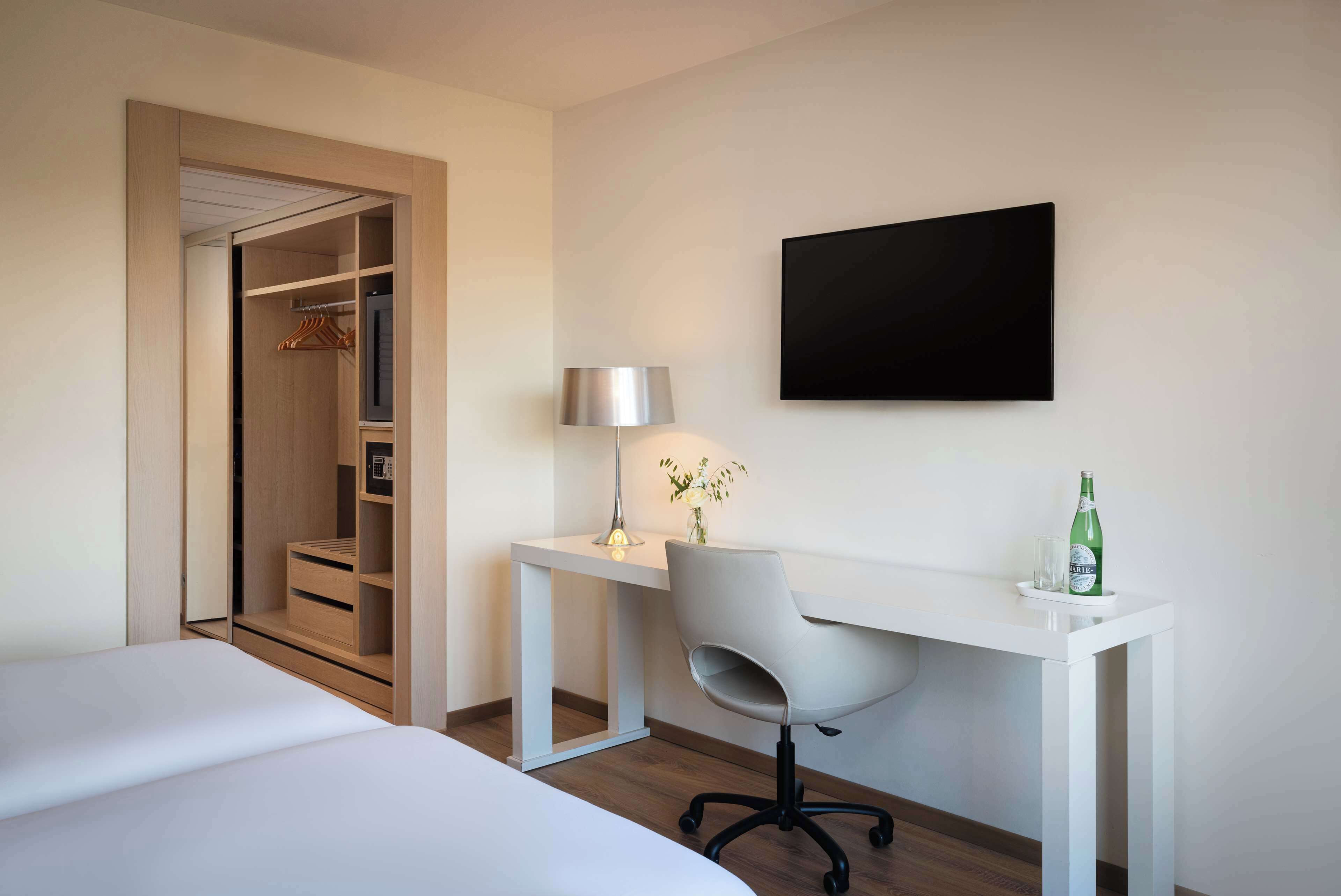 A hotel room with a bed, a desk with a chair, a TV, a lamp, and a bottle of water.