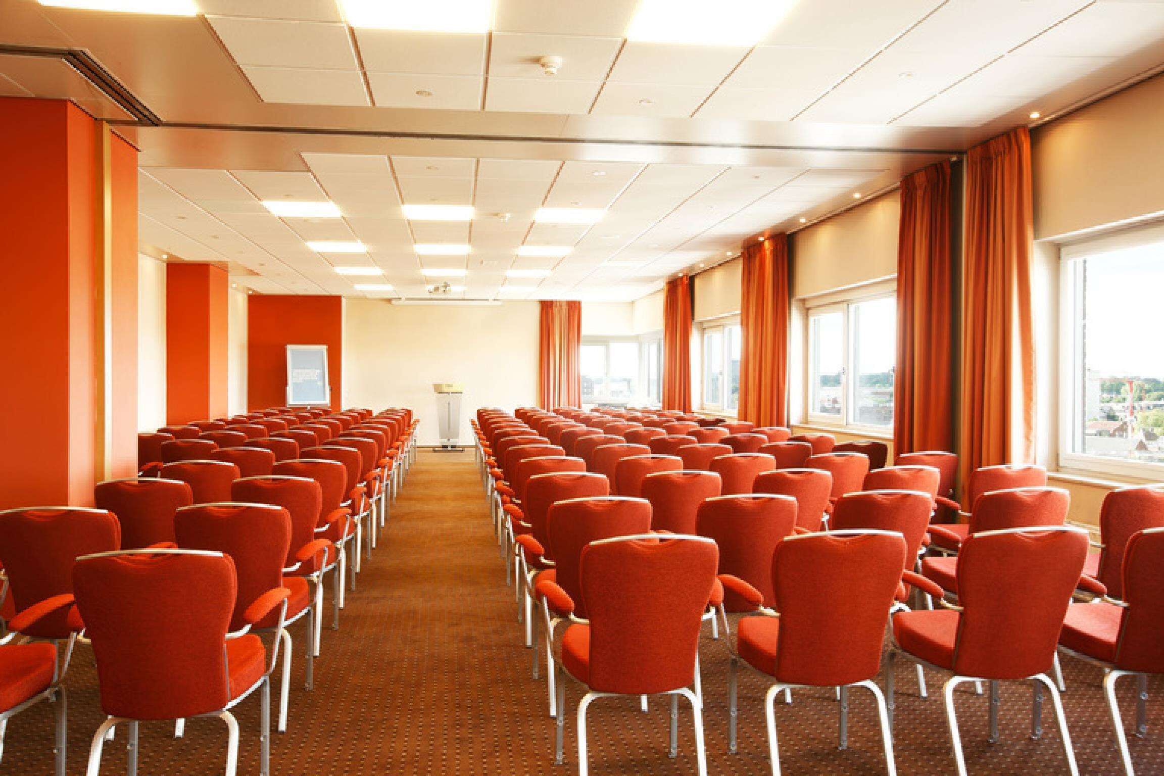 A conference room with rows of red chairs, a carpeted floor, and large windows with curtains.