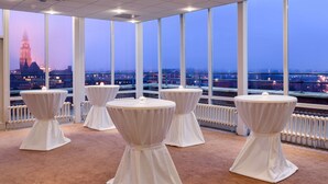 Una habitación con amplios ventanales, mesas blancas con tapas redondas y vistas a un paisaje urbano al atardecer.