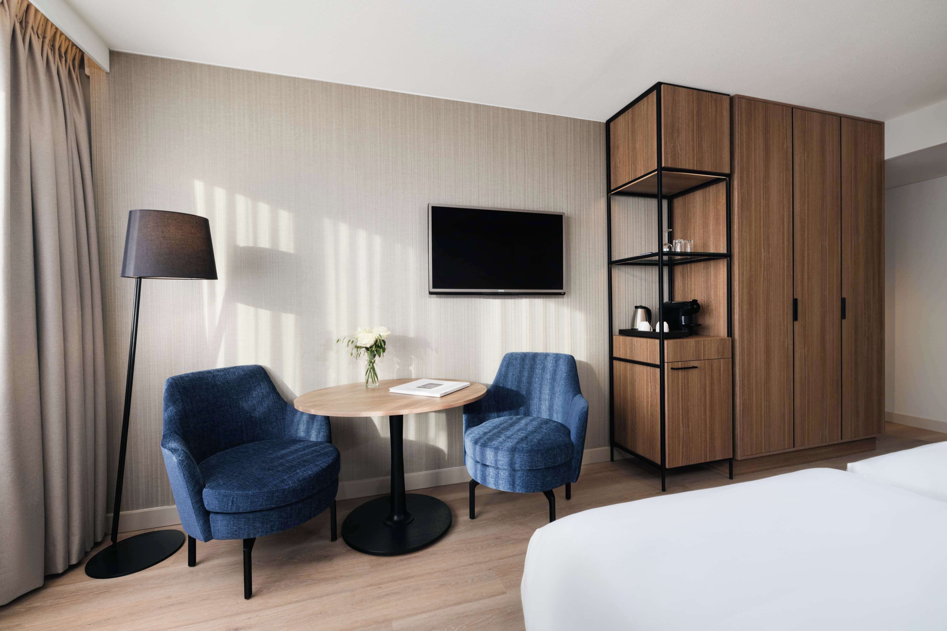 A hotel room with a bed, a TV mounted on the wall, a wooden wardrobe, two blue armchairs, a round table with a vase of flowers, and a floor lamp.