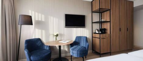 A hotel room with a bed, a TV mounted on the wall, a wooden wardrobe, two blue armchairs, a round table with a vase of flowers, and a floor lamp.