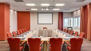 A conference room with a long table, red chairs, a projector screen, and a large window with curtains.