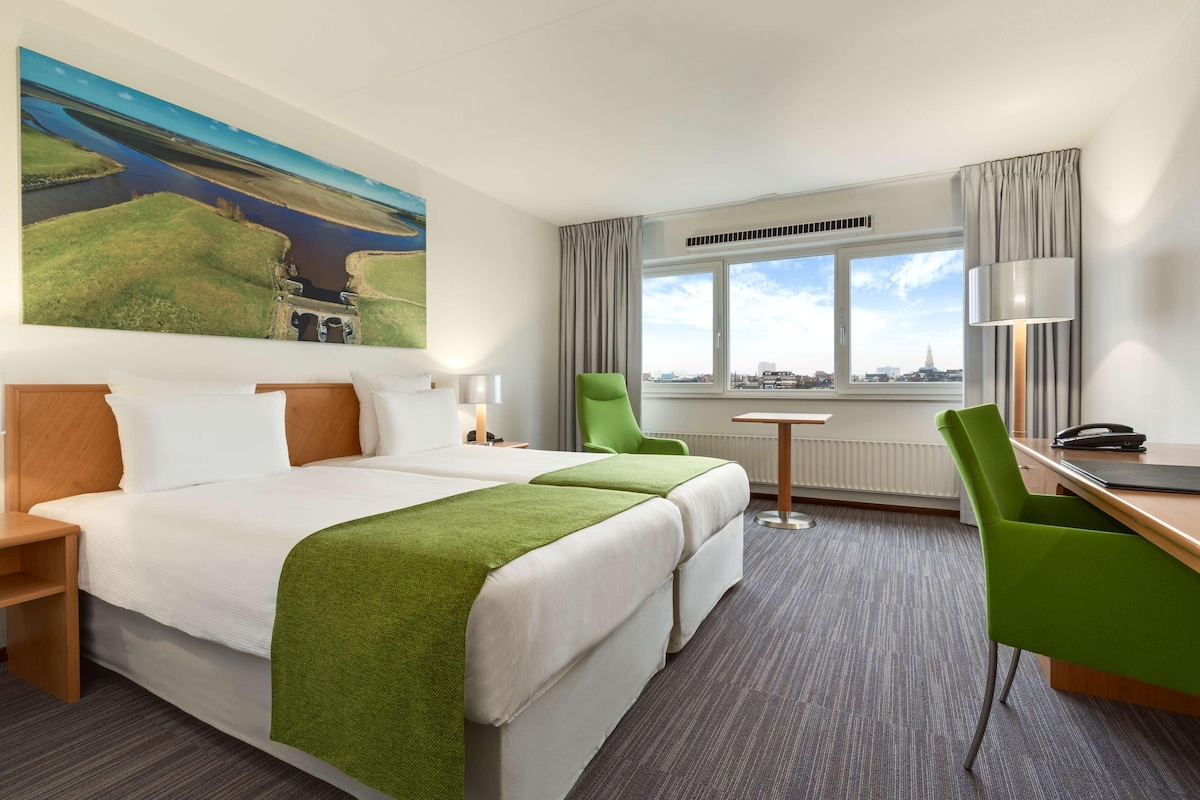 Habitación de hotel con una cama grande, dos sillas verdes, un escritorio y vista a un paisaje urbano por la ventana.