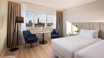A hotel room with a large bed, two armchairs, a small table, and a view of a cityscape.