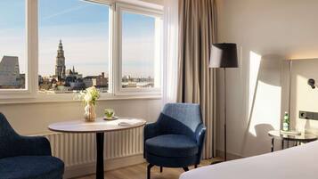 A hotel room with a large window offering a city view, a round table with a vase of flowers, two blue armchairs, a bed, and a floor lamp.