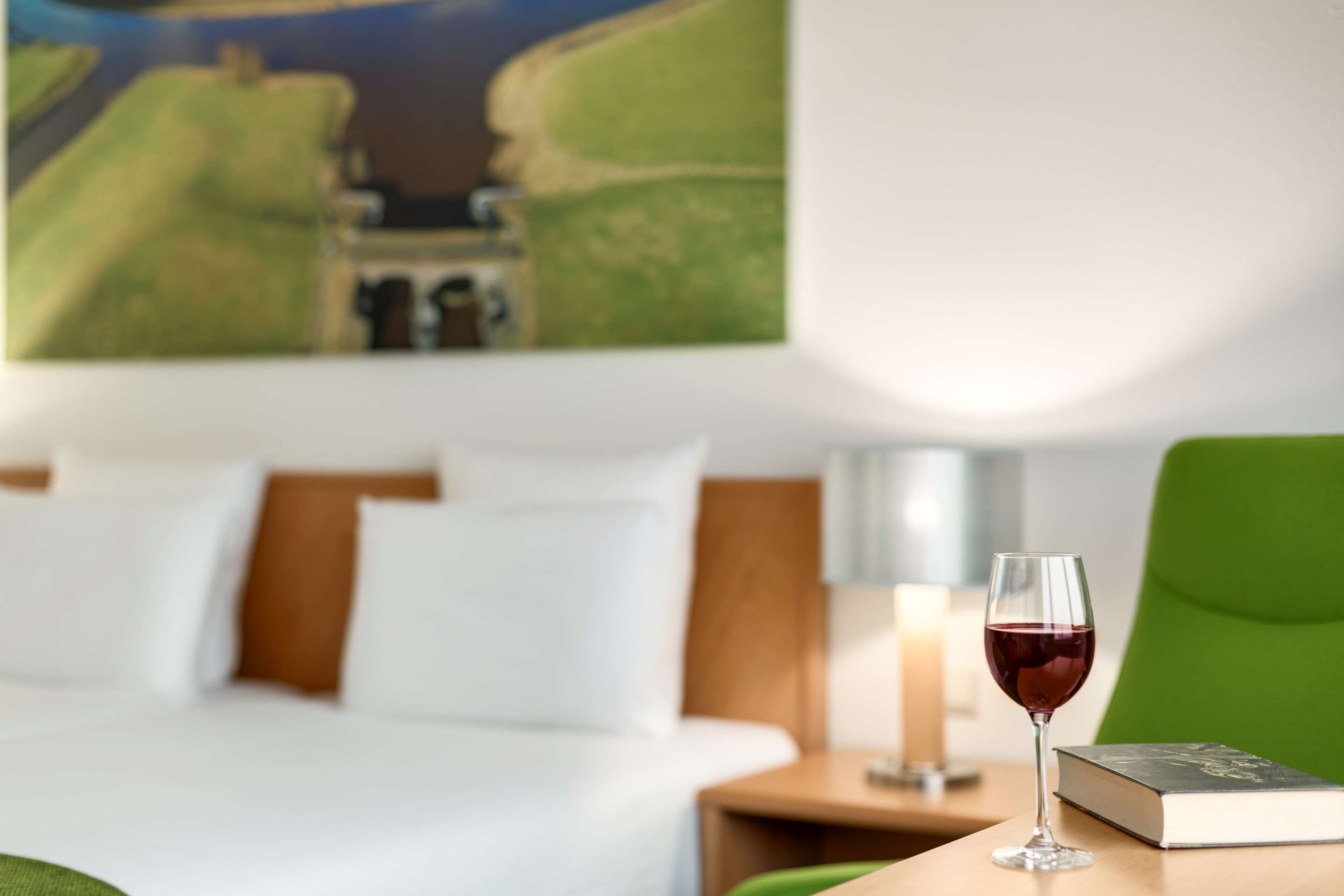 A hotel room with a bed, a green chair, a glass of red wine, and a book on a small table.