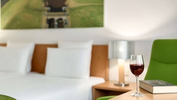 A hotel room with a bed, a green chair, a glass of red wine, and a book on a small table.