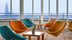 Habitación moderna con ventanas amplias, sillas naranja y aguamarina, una mesita y vistas a una torre lejana.