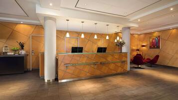 A reception desk with a modern design, featuring a geometric patterned wall, a glass partition, and a metal railing. There are three pendant lights above the desk, and a painting is visible on the wall to the right.