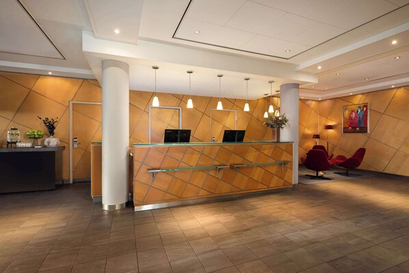 A reception desk with a modern design, featuring a geometric patterned wall, a glass partition, and a metal railing. There are three pendant lights above the desk, and a painting is visible on the wall to the right.