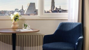 Una habitación con vistas a un paisaje urbano, un sillón azul, una mesita con un florero y un libro.