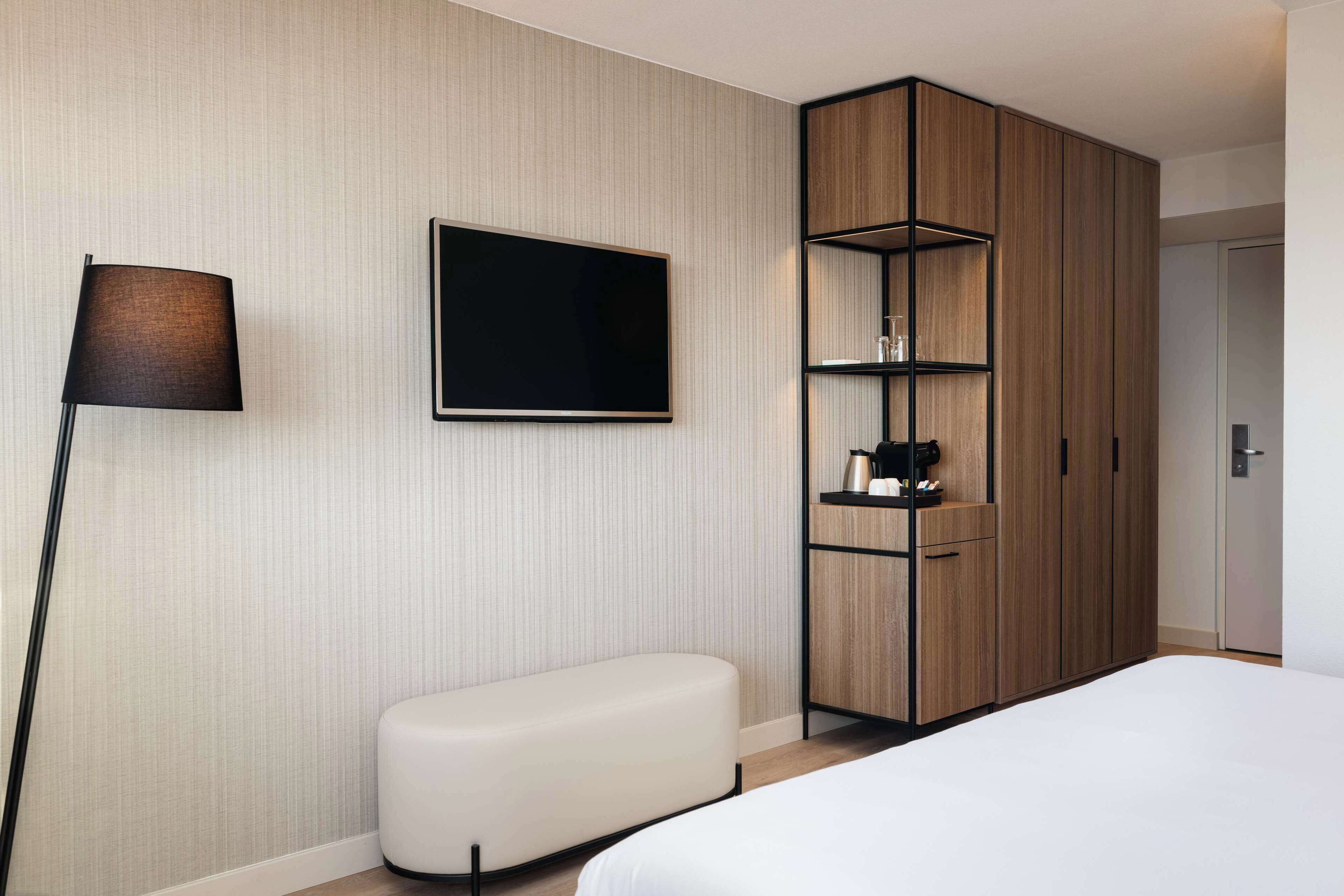 Een moderne hotelkamer met een groot bed, een flatscreen televisie aan de muur, een houten garderobe met planken, en een klein wit bankje.