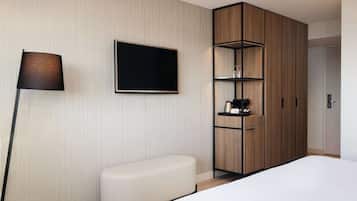 Een moderne hotelkamer met een groot bed, een flatscreen televisie aan de muur, een houten garderobe met planken, en een klein wit bankje.