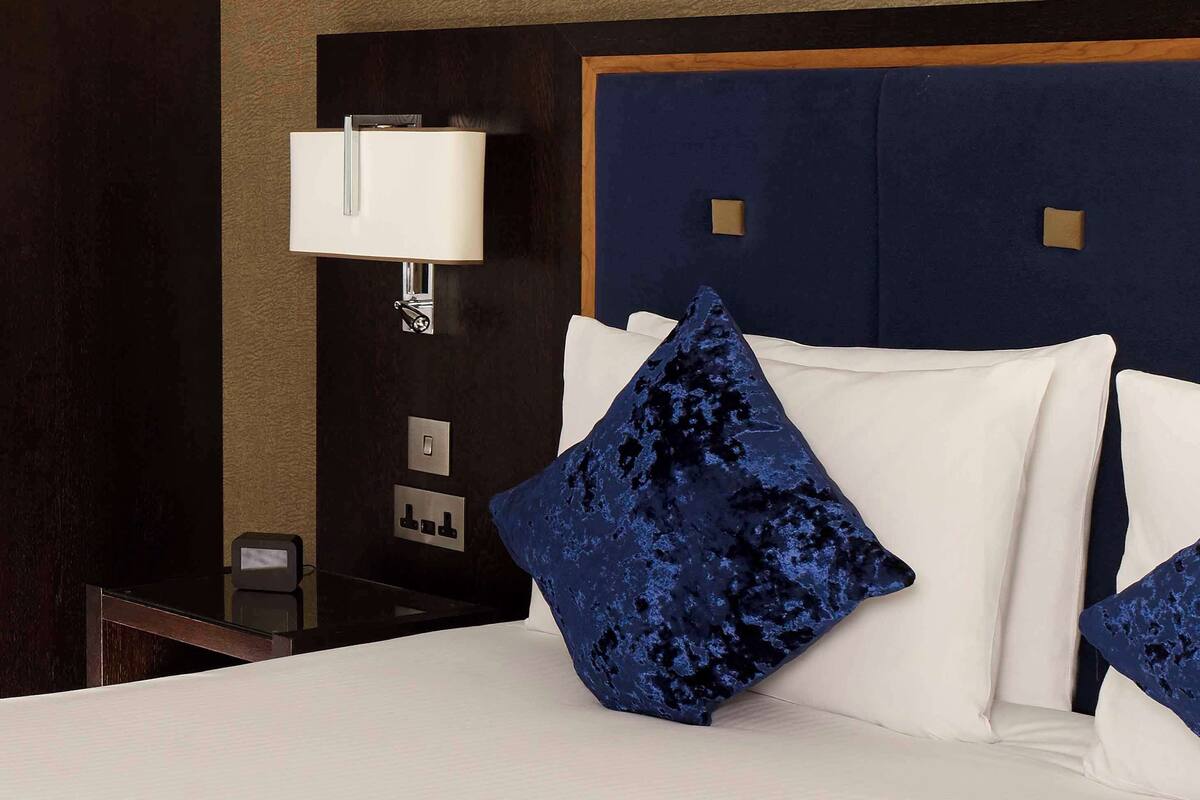 Habitación de hotel con cama, ropa de cama blanca, almohada azul, lámpara fijada a la pared y cabecero de madera.