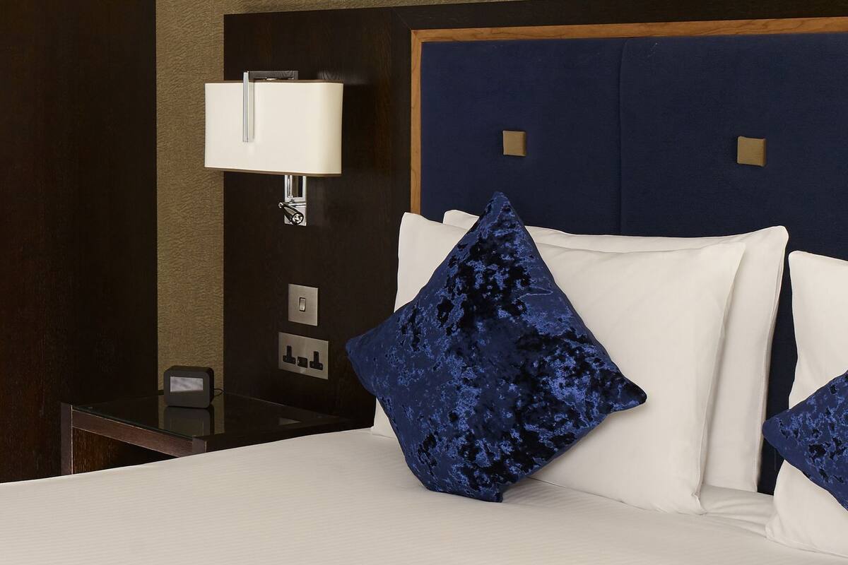A hotel room with a bed, a nightstand, a lamp, and a wall-mounted control panel.