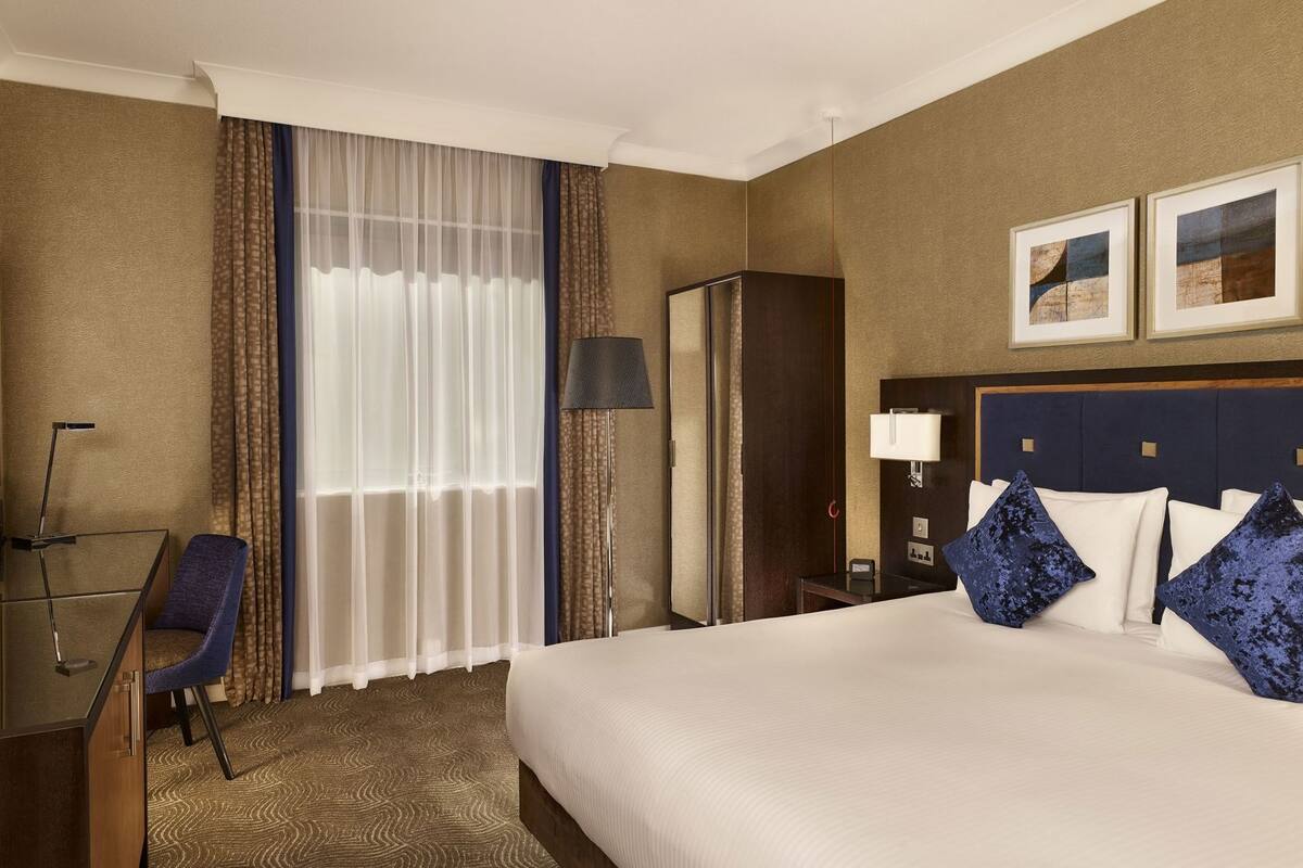 A hotel room with a bed, a desk, a chair, a lamp, and a mirror.