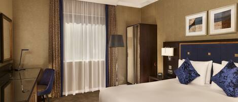 A hotel room with a large bed, a desk, a chair, a lamp, a wardrobe, and a mirror.