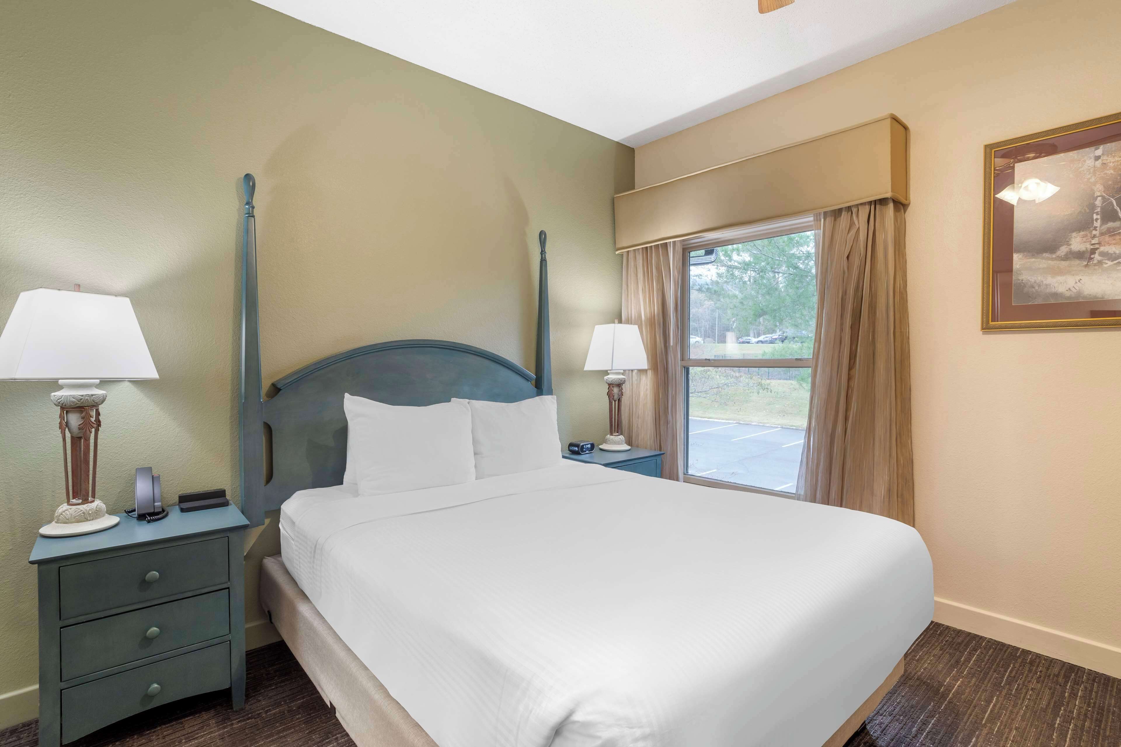 A hotel room with a bed, two bedside tables with lamps, a window with curtains, and a painting on the wall.