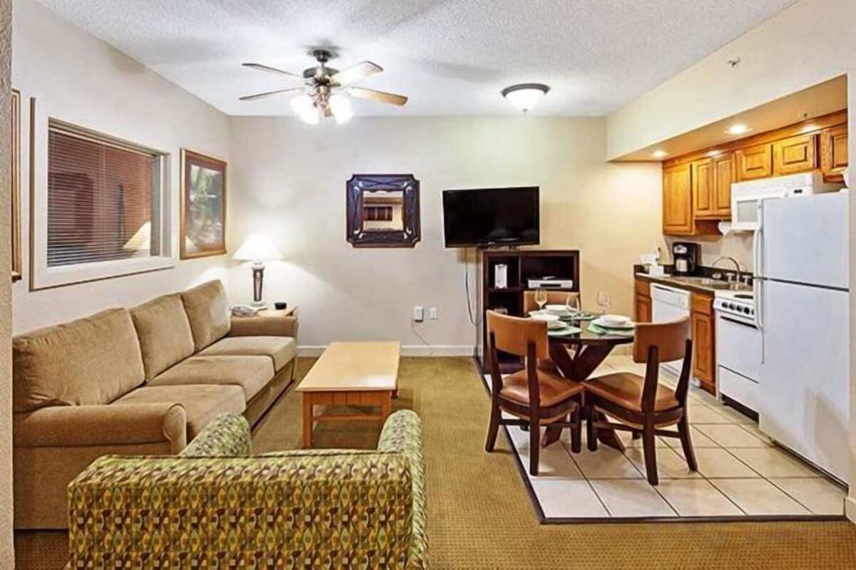 A compact living space with a sofa, a patterned armchair, a dining table with chairs, a television, and a kitchenette with a refrigerator and stove.