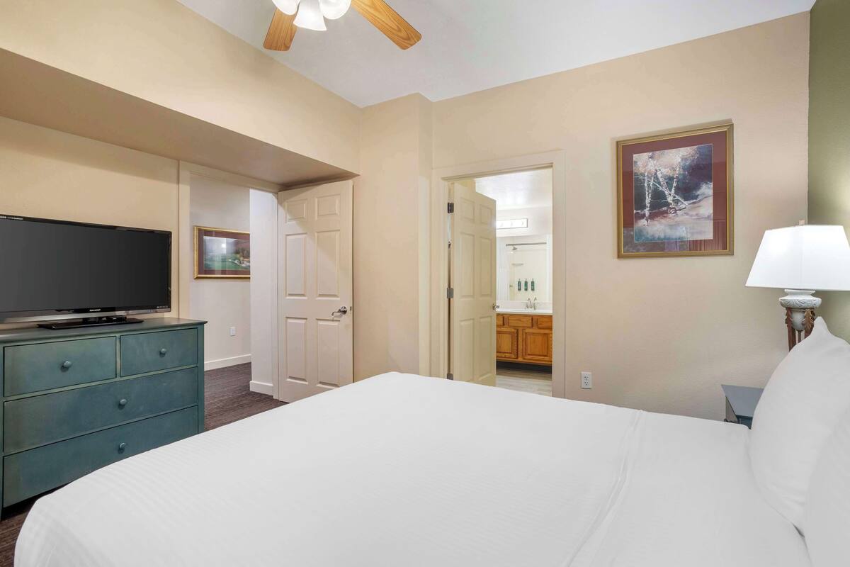 A hotel room with a bed, a dresser, a television, a painting, and a bathroom visible through an open door.